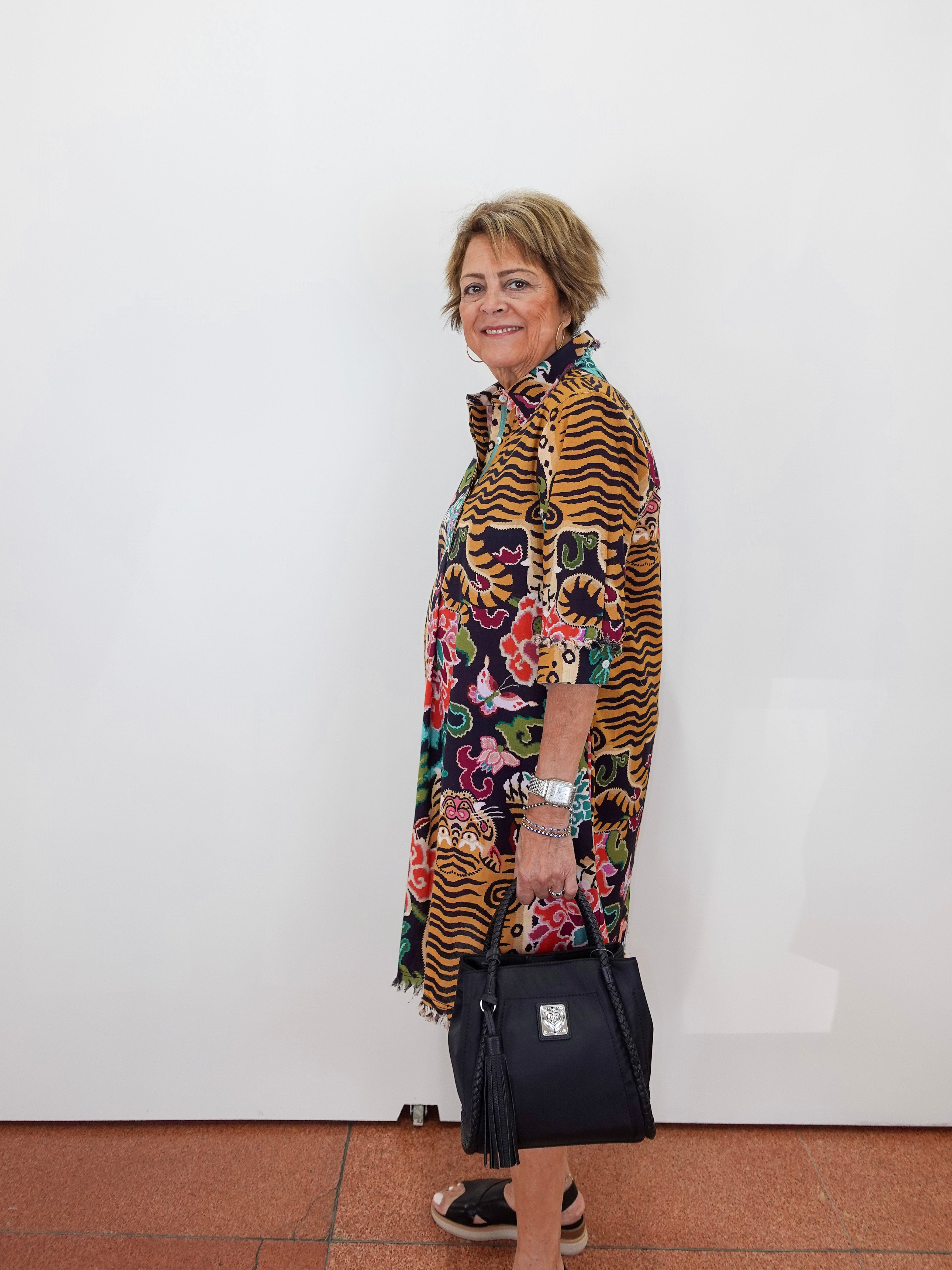 A woman stands in profile against a plain white wall, wearing a long-sleeved, open-front tunic dress with a bold multicolor print featuring tiger stripes, floral motifs, and colorful abstract shapes. She is holding a black handbag and wearing black sandals.