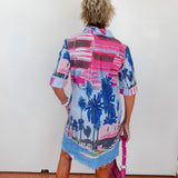 Person wearing a colorful palm tree patterned shirt over a dress, holding a pink bag.