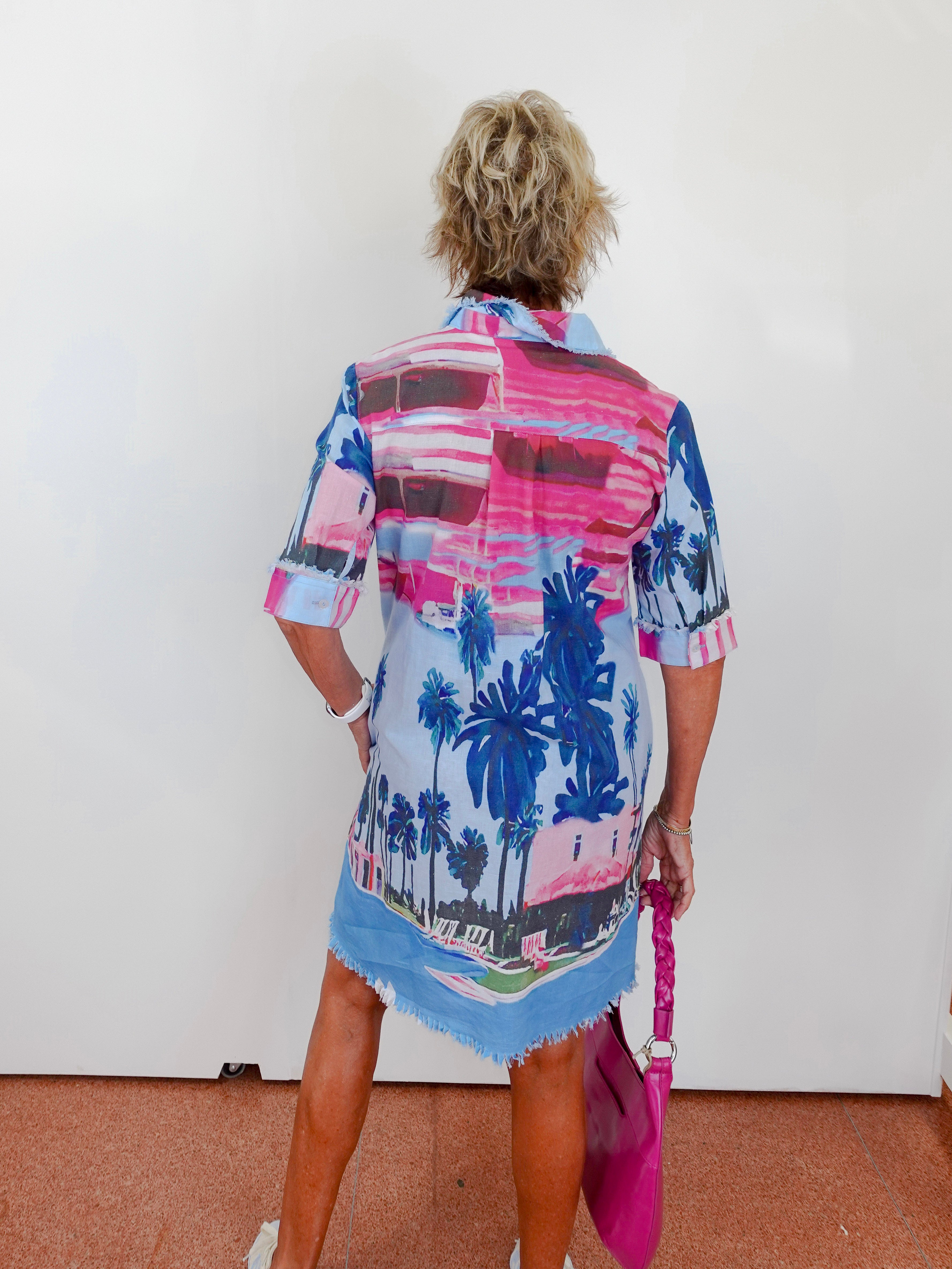 Person wearing a colorful palm tree patterned shirt over a dress, holding a pink bag.