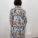 A person viewed from behind wearing a short-sleeve button-up shirt dress with a dense multicolor nature print featuring insects, spiders, and plants on a white base, standing against a plain white wall.
