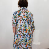 A person viewed from behind wearing a short-sleeve button-up shirt dress with a dense multicolor nature print featuring insects, spiders, and plants on a white base, standing against a plain white wall.
