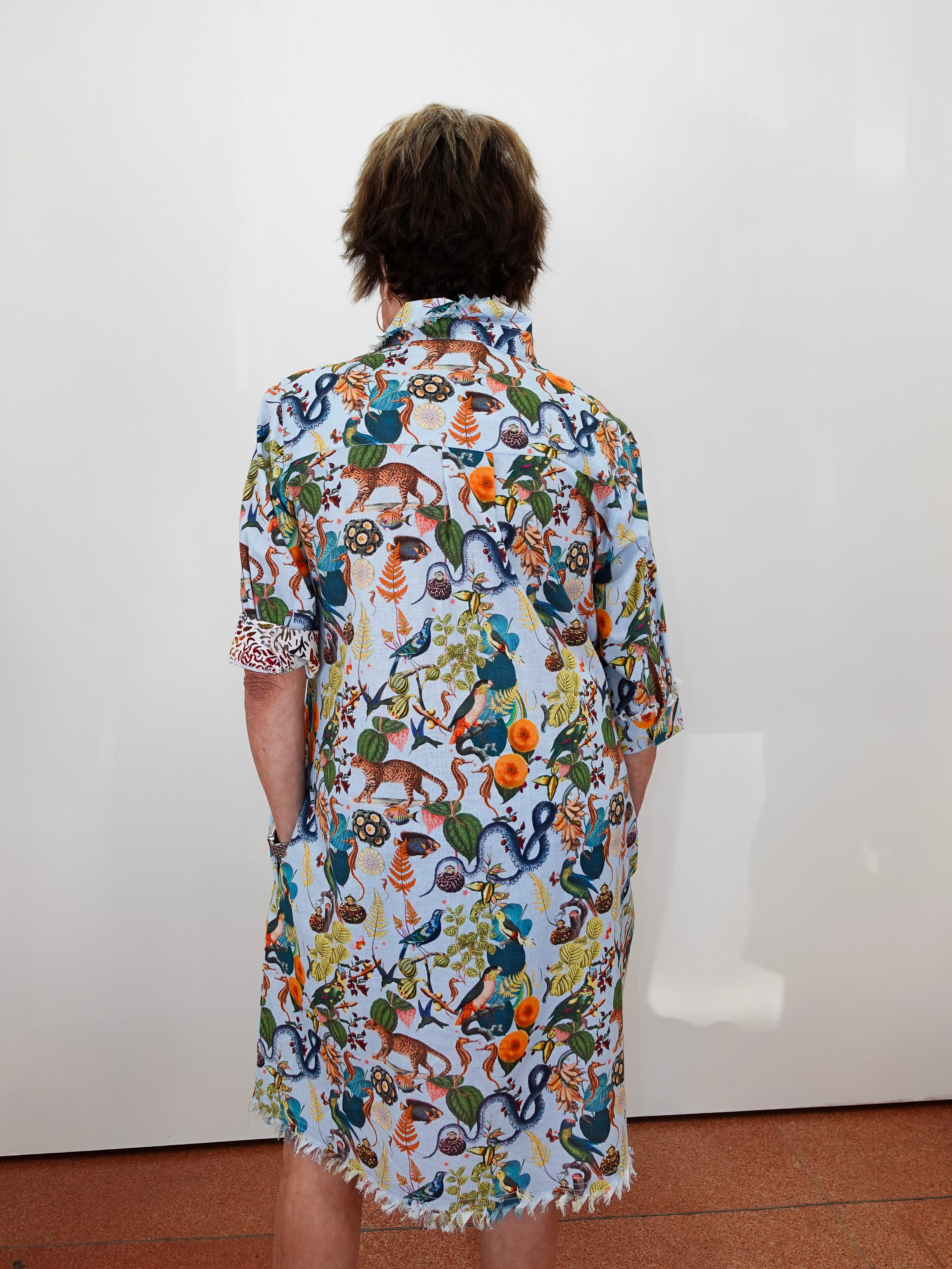 A person viewed from behind wearing a short-sleeve button-up shirt dress with a dense multicolor nature print featuring insects, spiders, and plants on a white base, standing against a plain white wall.