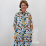 A woman with short brown hair stands against a white wall wearing a white button-up shirt dress covered in a colorful, dense cartoon-style print featuring animals and foliage.