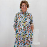 A woman with short brown hair stands against a white wall wearing a white button-up shirt dress covered in a colorful, dense cartoon-style print featuring animals and foliage.