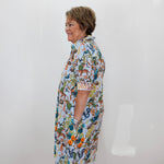 An older woman with short brown hair stands in profile wearing a short-sleeve button-up shirt dress with a colorful, busy botanical and animal print on a white base. She is standing against a plain white wall with a terracotta tiled floor visible at the bottom.