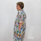 An older woman with short brown hair stands in profile wearing a short-sleeve button-up shirt dress with a colorful, busy botanical and animal print on a white base. She is standing against a plain white wall with a terracotta tiled floor visible at the bottom.