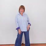 Woman wearing a blue and white striped shirt and blue jeans standing against a white wall.