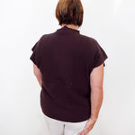 A woman viewed from behind wearing a dark plum ribbed short-sleeve mock neck top and white trousers, standing against a plain white wall.