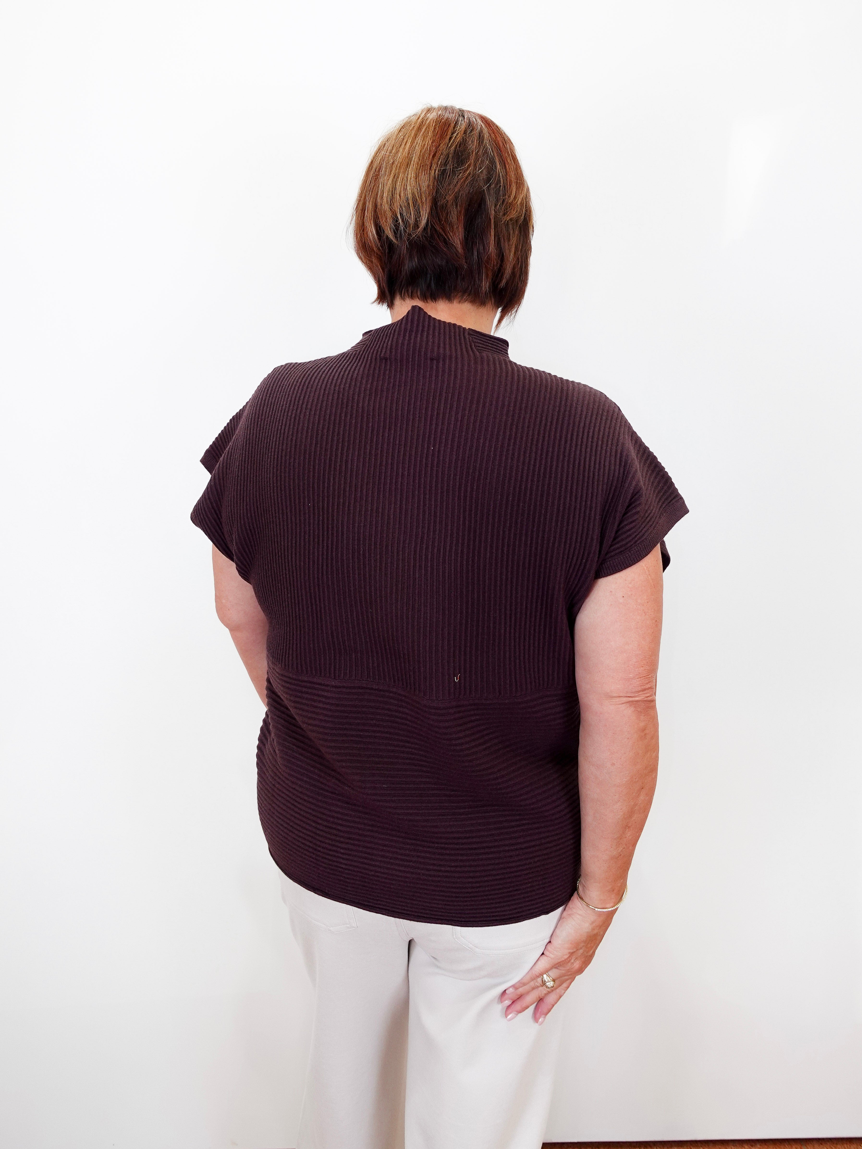 A woman viewed from behind wearing a dark plum ribbed short-sleeve mock neck top and white trousers, standing against a plain white wall.
