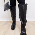 Person wearing black knee-high boots and holding a matching handbag on a light wooden floor.