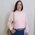 Woman wearing a pink sweater and blue jeans holding a green bag against a white wall.