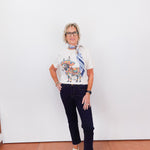 Person wearing a white t-shirt with a graphic design and dark jeans against a white wall.