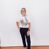 Person wearing a white t-shirt with a graphic design and dark jeans against a white wall.