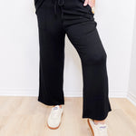 Person wearing a black outfit with wide-leg pants and white sneakers on a light wooden floor.