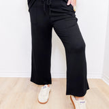 Person wearing a black outfit with wide-leg pants and white sneakers on a light wooden floor.