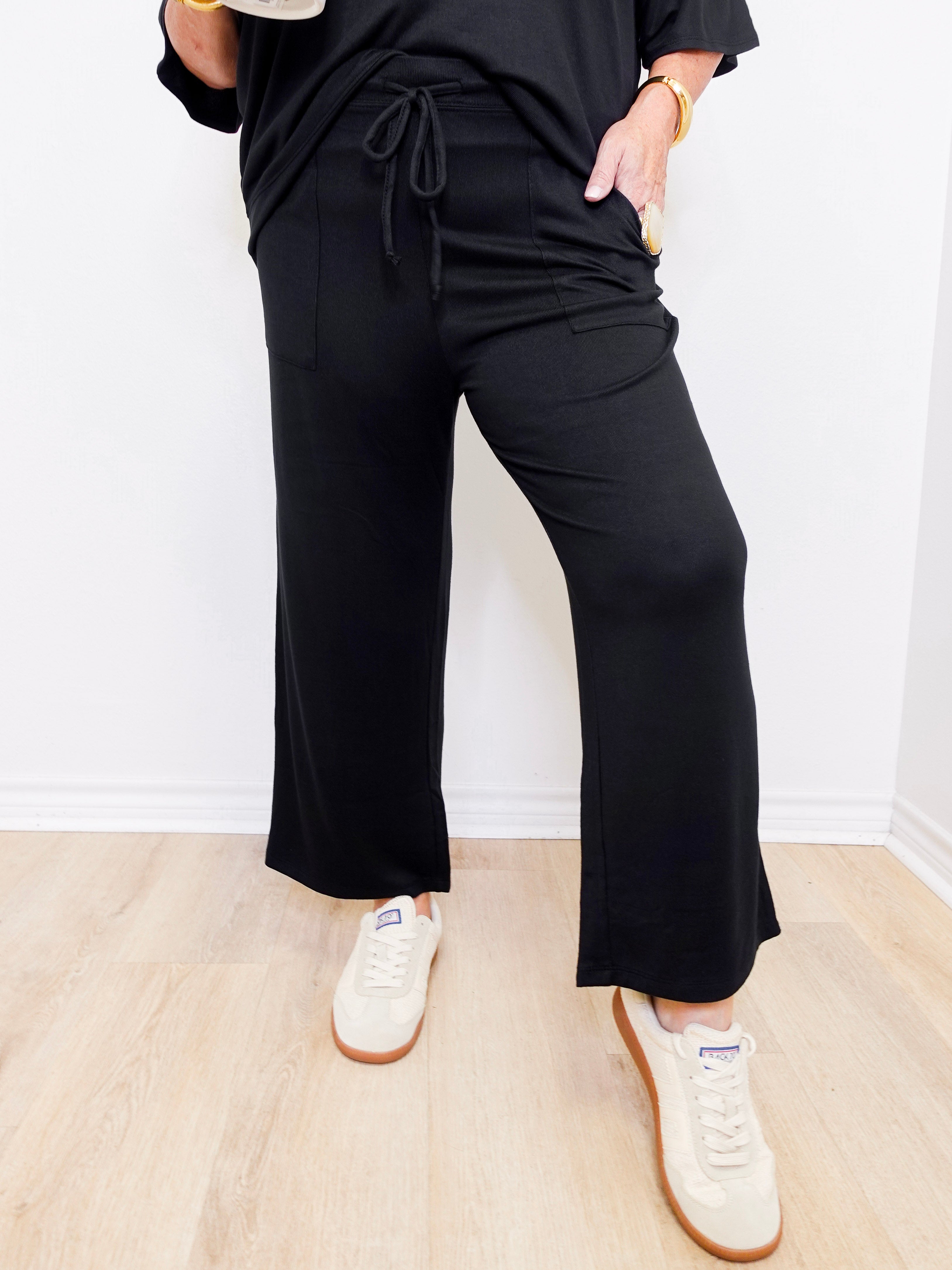 Person wearing a black outfit with wide-leg pants and white sneakers on a light wooden floor.