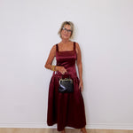 Woman in a burgundy dress holding a black handbag against a white wall.