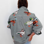 A person viewed from behind wearing a black and white gingham button-up shirt with colorful embroidered ducks across the fabric, paired with light blue jeans against a plain white wall.