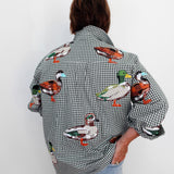 A person viewed from behind wearing a black and white gingham button-up shirt with colorful embroidered ducks across the fabric, paired with light blue jeans against a plain white wall.