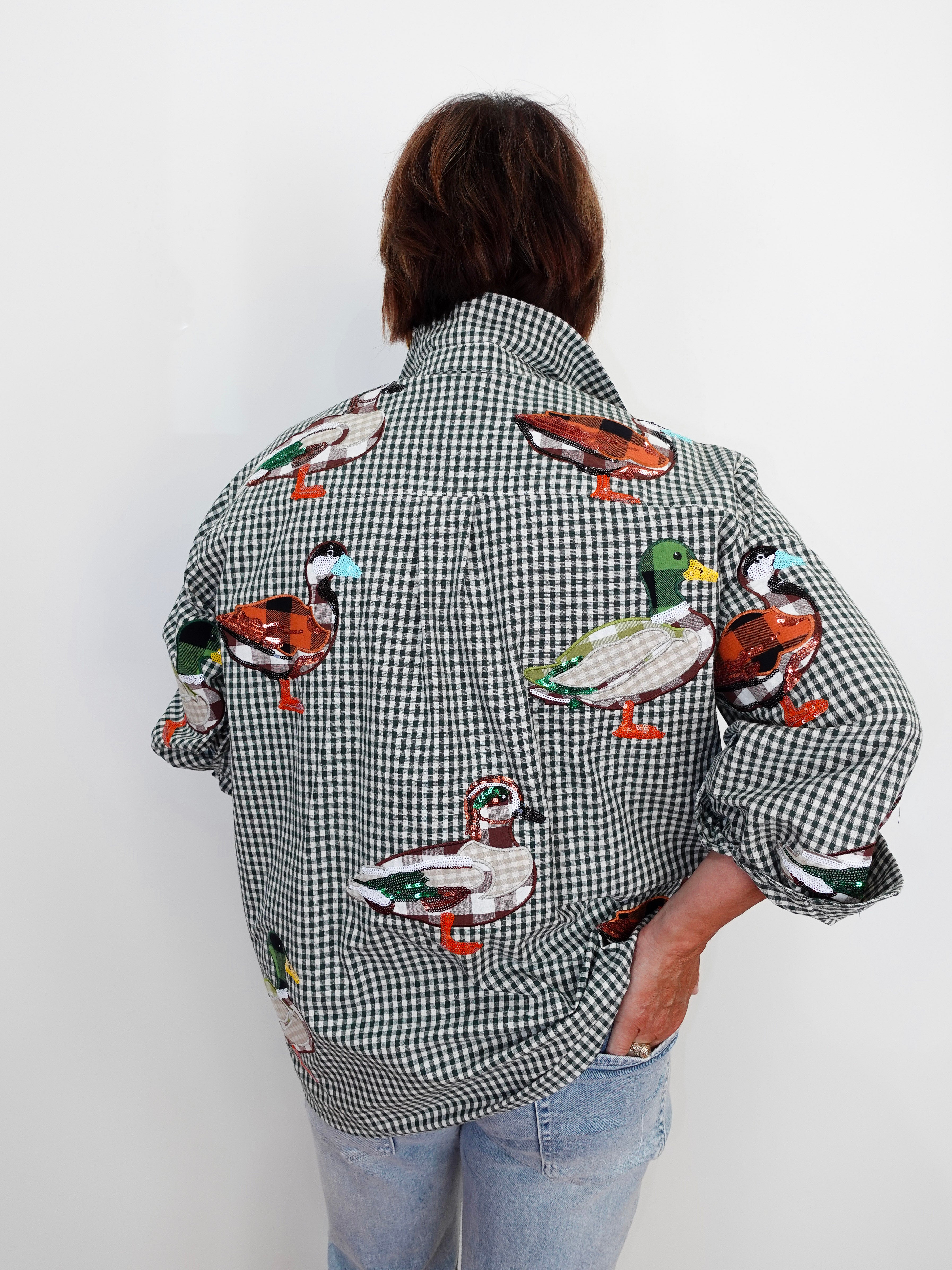 A person viewed from behind wearing a black and white gingham button-up shirt with colorful embroidered ducks across the fabric, paired with light blue jeans against a plain white wall.