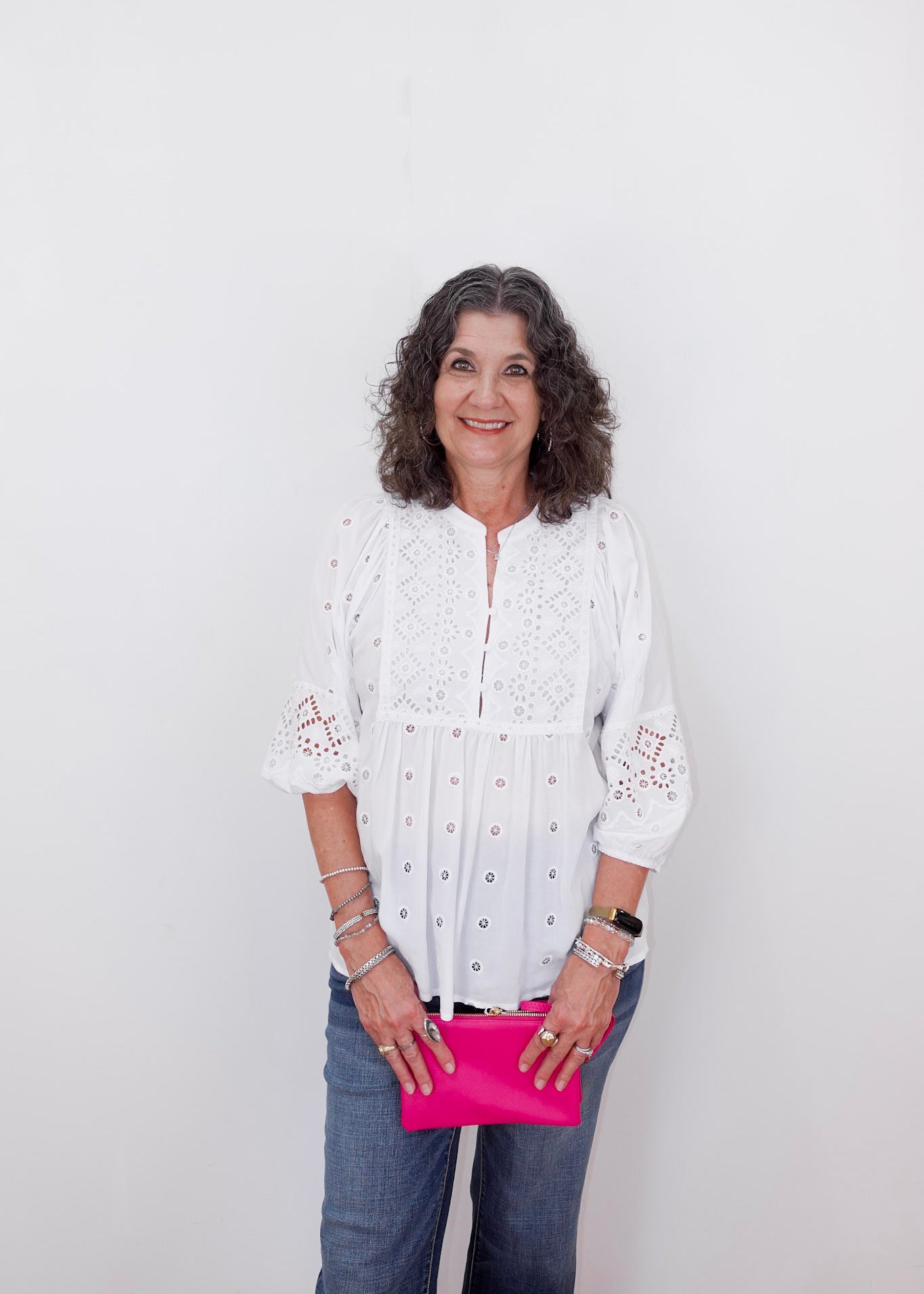 Woman wearing a white blouse with lace details and blue jeans, holding a pink clutch against a white background.