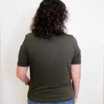 Person wearing an olive green t-shirt and blue jeans standing against a white wall.