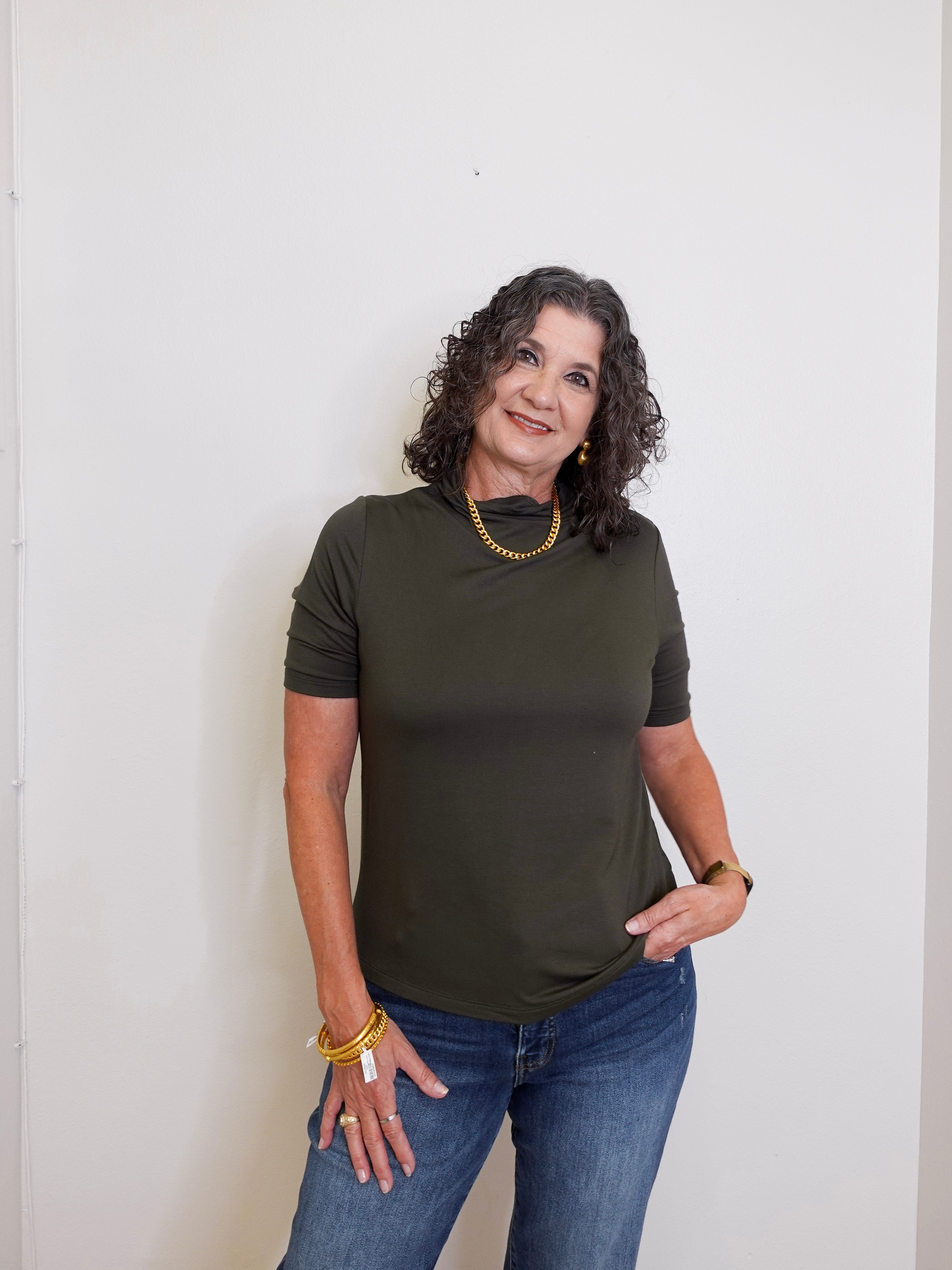 Woman wearing an olive green t-shirt and blue jeans against a white wall