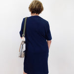 A woman viewed from behind wearing a navy blue short-sleeve dress and carrying a clear shoulder bag with a striped strap, standing against a plain white wall.