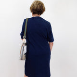 A woman viewed from behind wearing a navy blue short-sleeve dress and carrying a clear shoulder bag with a striped strap, standing against a plain white wall.