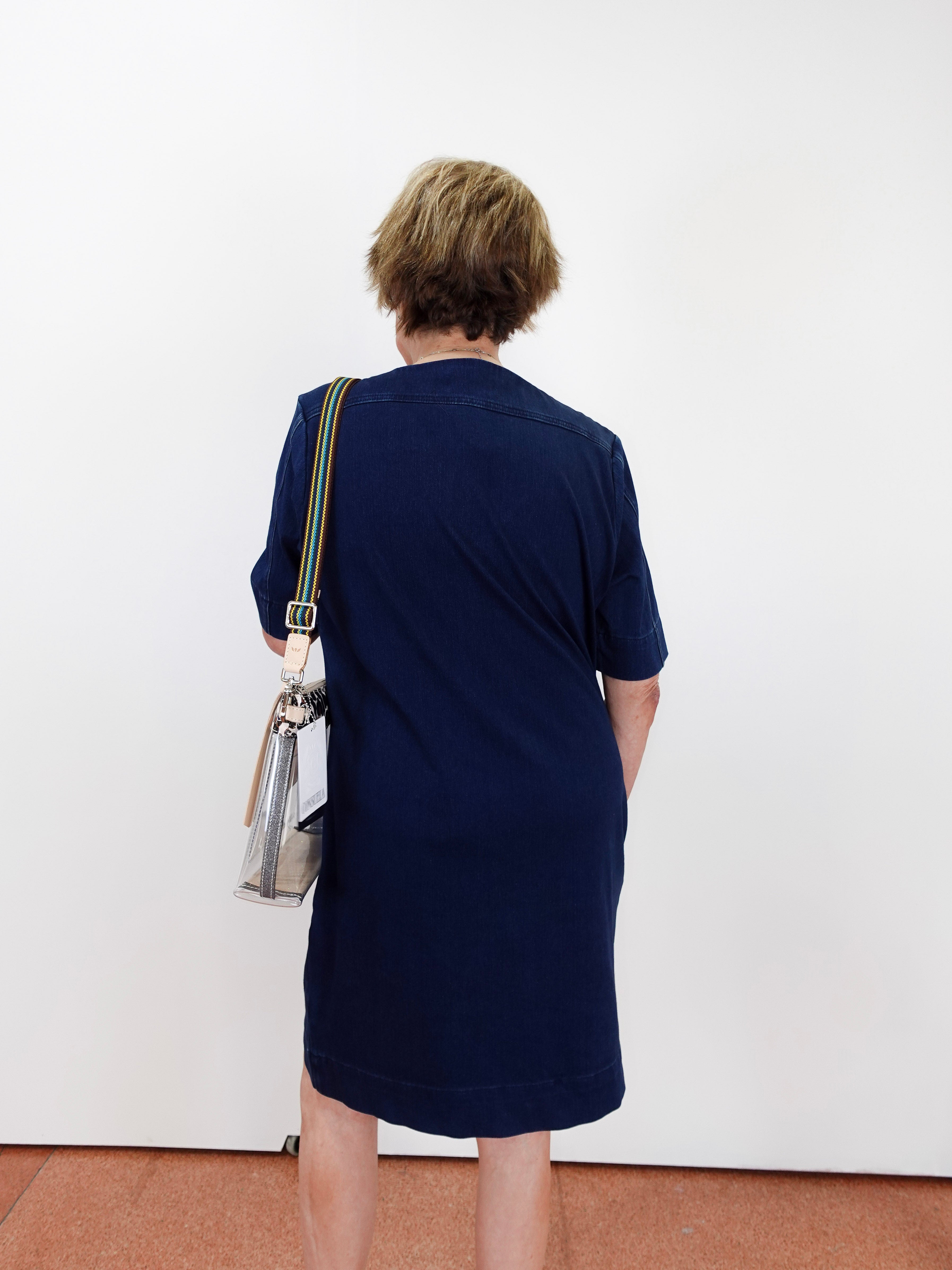 A woman viewed from behind wearing a navy blue short-sleeve dress and carrying a clear shoulder bag with a striped strap, standing against a plain white wall.