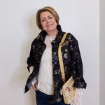 Woman wearing a black floral jacket with a white top and blue jeans, holding a beige handbag against a white background.