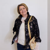 Woman wearing a black floral jacket with a white top and blue jeans, holding a beige handbag against a white background.
