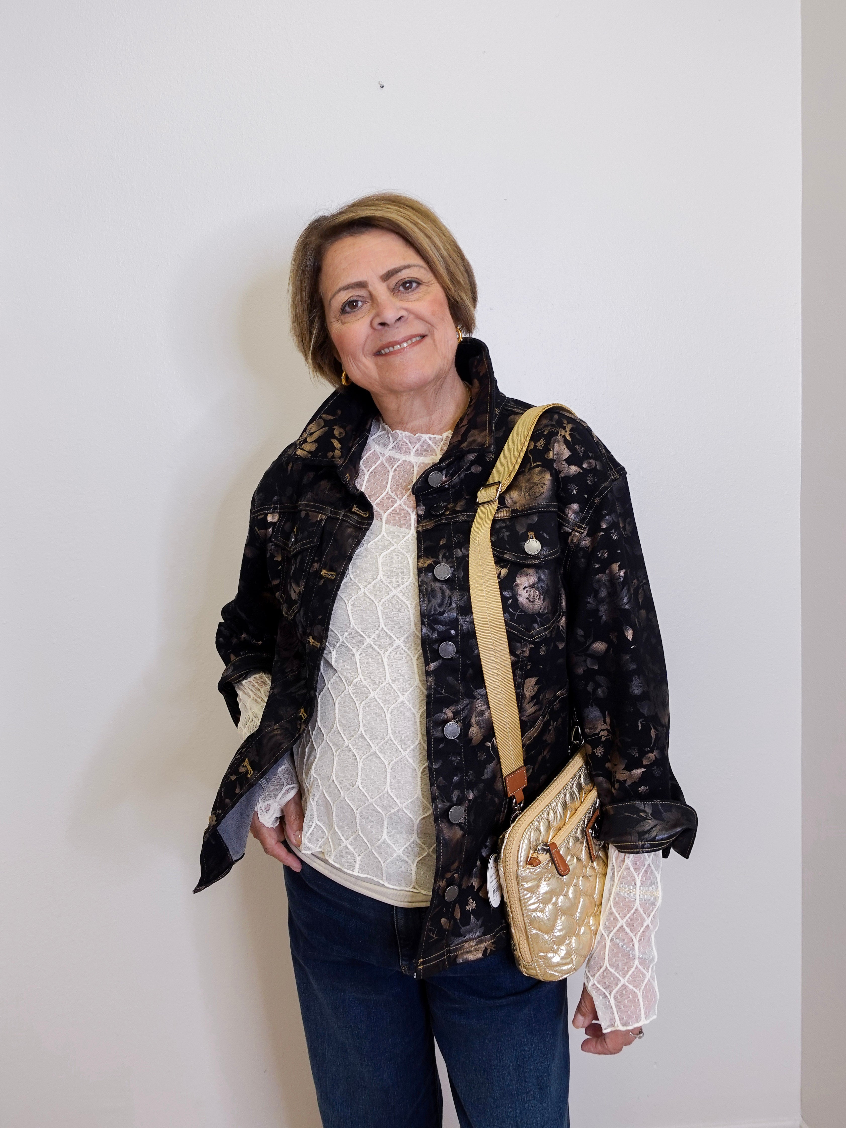 Woman wearing a black floral jacket with a white top and blue jeans, holding a beige handbag against a white background.