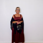 Woman in a burgundy dress with a black shawl holding a handbag against a white wall.