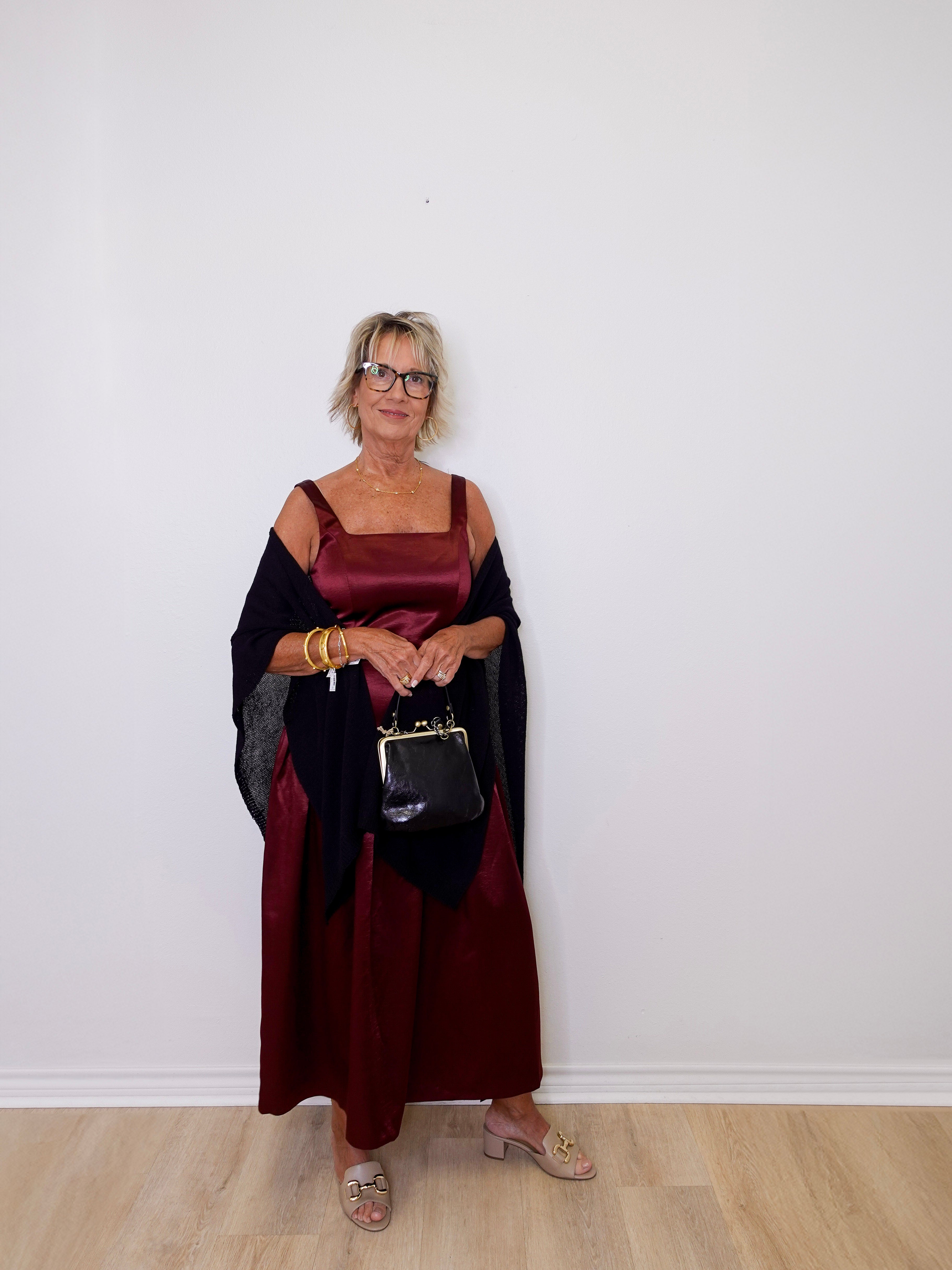 Woman in a burgundy dress with a black shawl holding a handbag against a white wall.