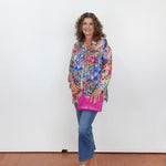 Woman wearing a colorful floral blouse and blue jeans, holding a pink bag against a white background.