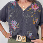 Woman wearing a dark gray blouse with floral and geometric embroidery, standing against a plain background.