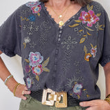 Woman wearing a dark gray blouse with floral and geometric embroidery, standing against a plain background.