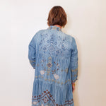 Person wearing a blue embroidered denim jacket against a white wall.