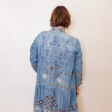 Person wearing a blue embroidered denim jacket against a white wall.