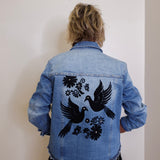 Person wearing a blue denim jacket with black bird and floral designs on the back against a plain background.