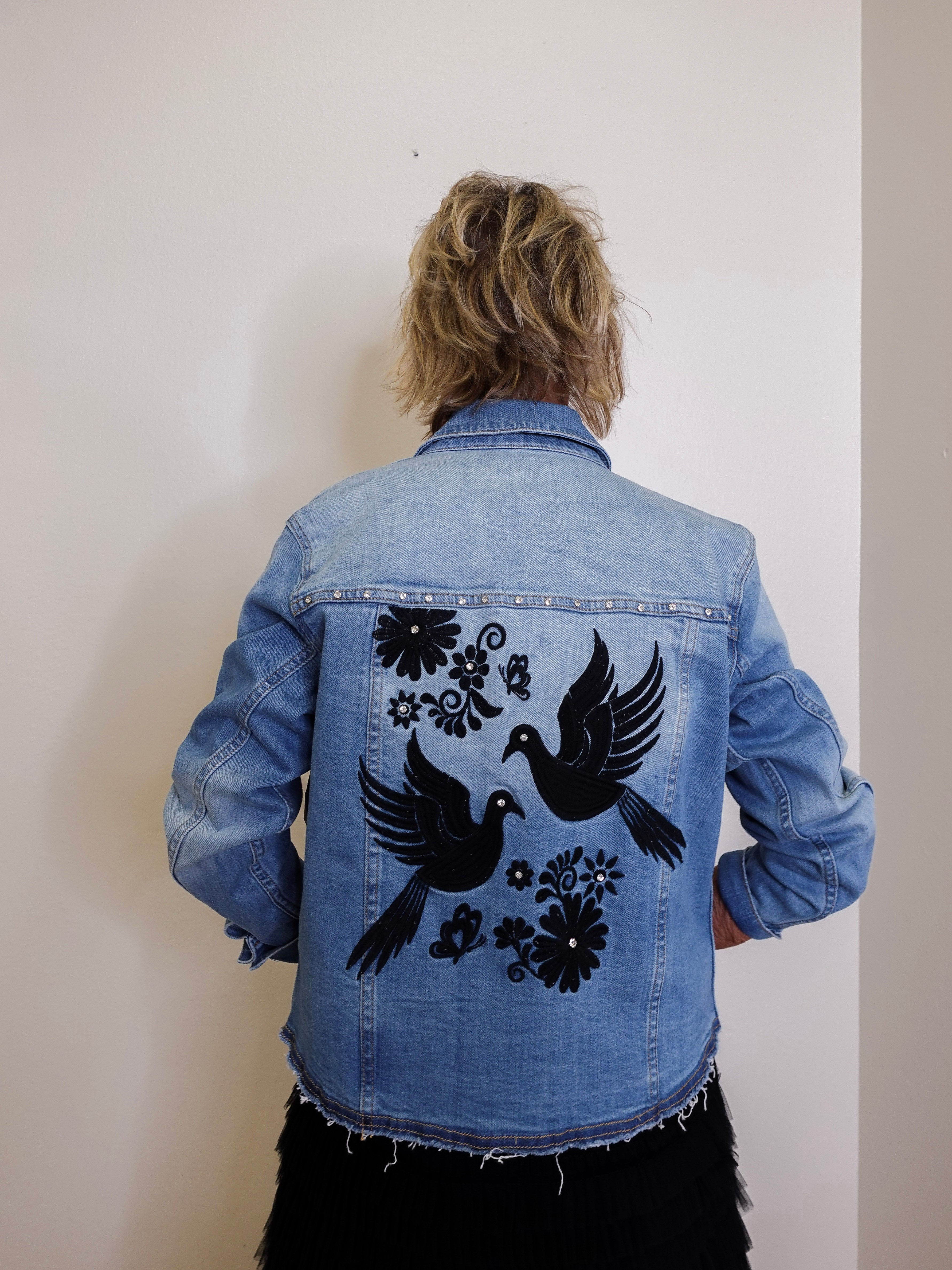 Person wearing a blue denim jacket with black bird and floral designs on the back against a plain background.