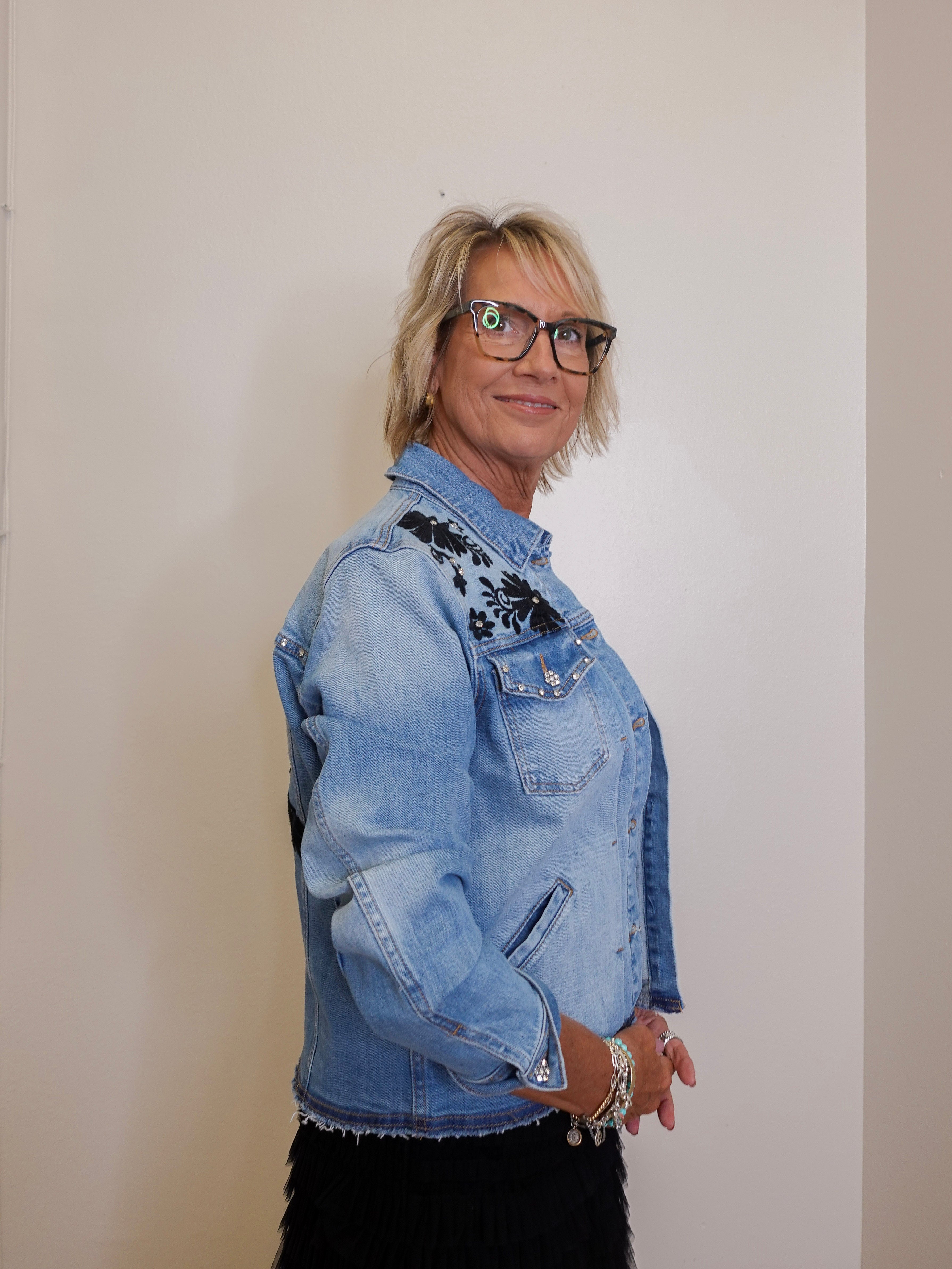 Person wearing a light blue denim jacket with floral embroidery on a plain background