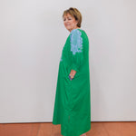 Woman wearing a green dress with blue lace details against a white wall.