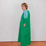 Woman wearing a green dress with blue lace details against a white wall.