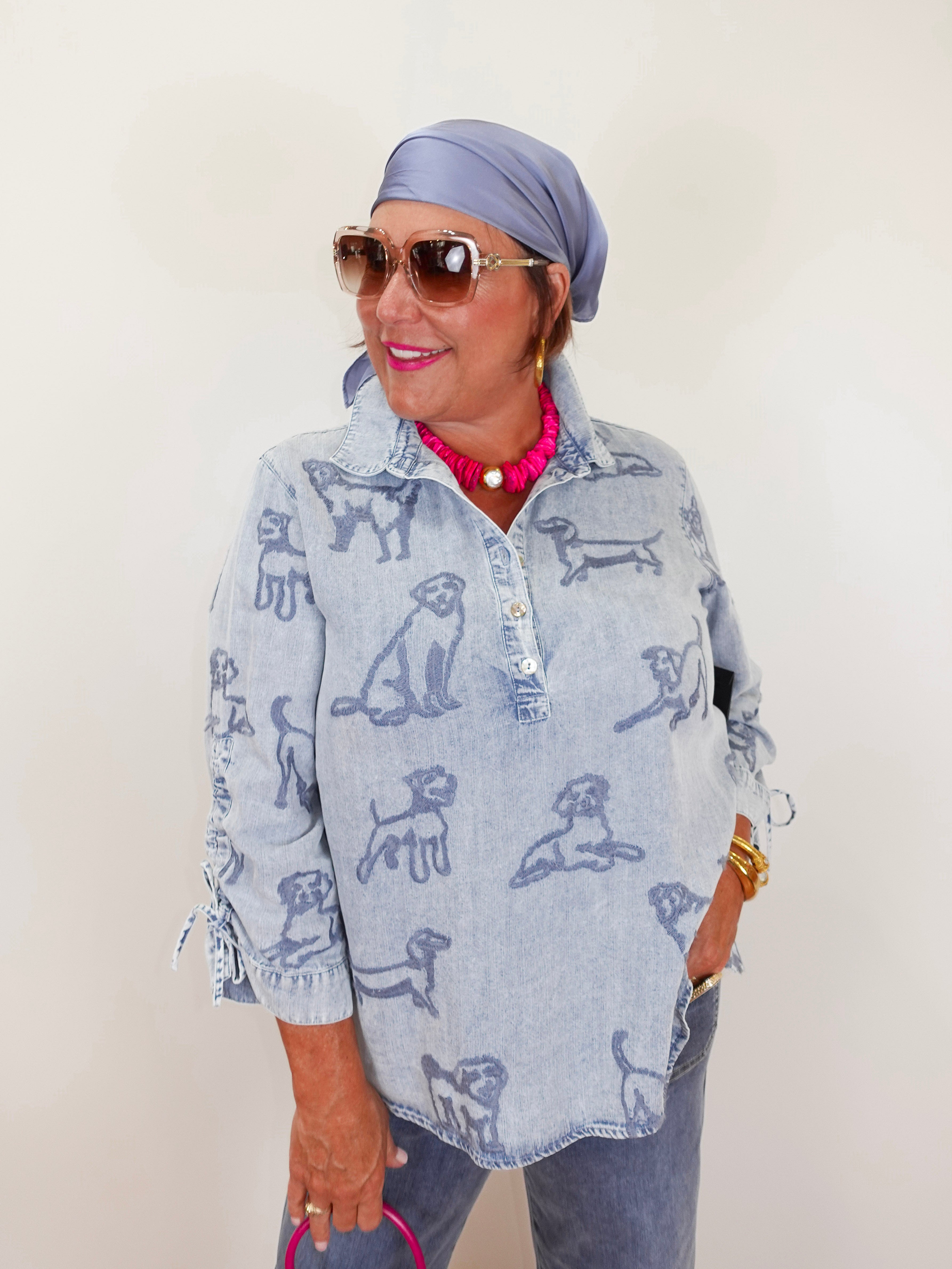 Woman wearing a light blue shirt with dog print, sunglasses, and headscarf against a plain background