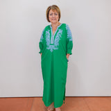 Woman wearing a green dress with blue embroidery against a white wall.