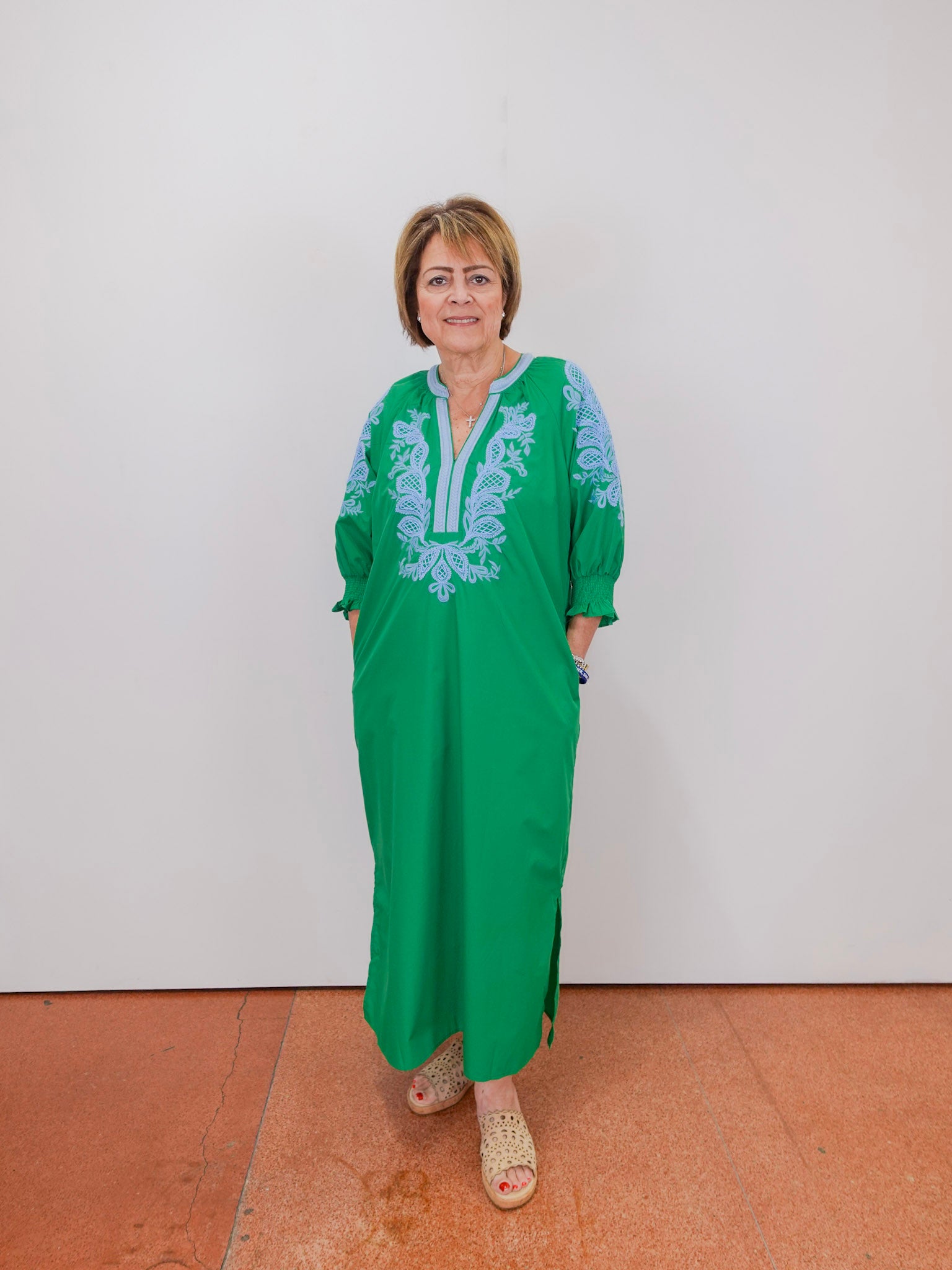 Woman wearing a green dress with blue embroidery against a white wall.