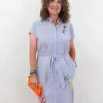 Woman wearing a light blue dress with floral embroidery, holding an orange clutch against a white background
