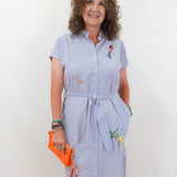 Woman wearing a light blue dress with floral embroidery, holding an orange clutch against a white background